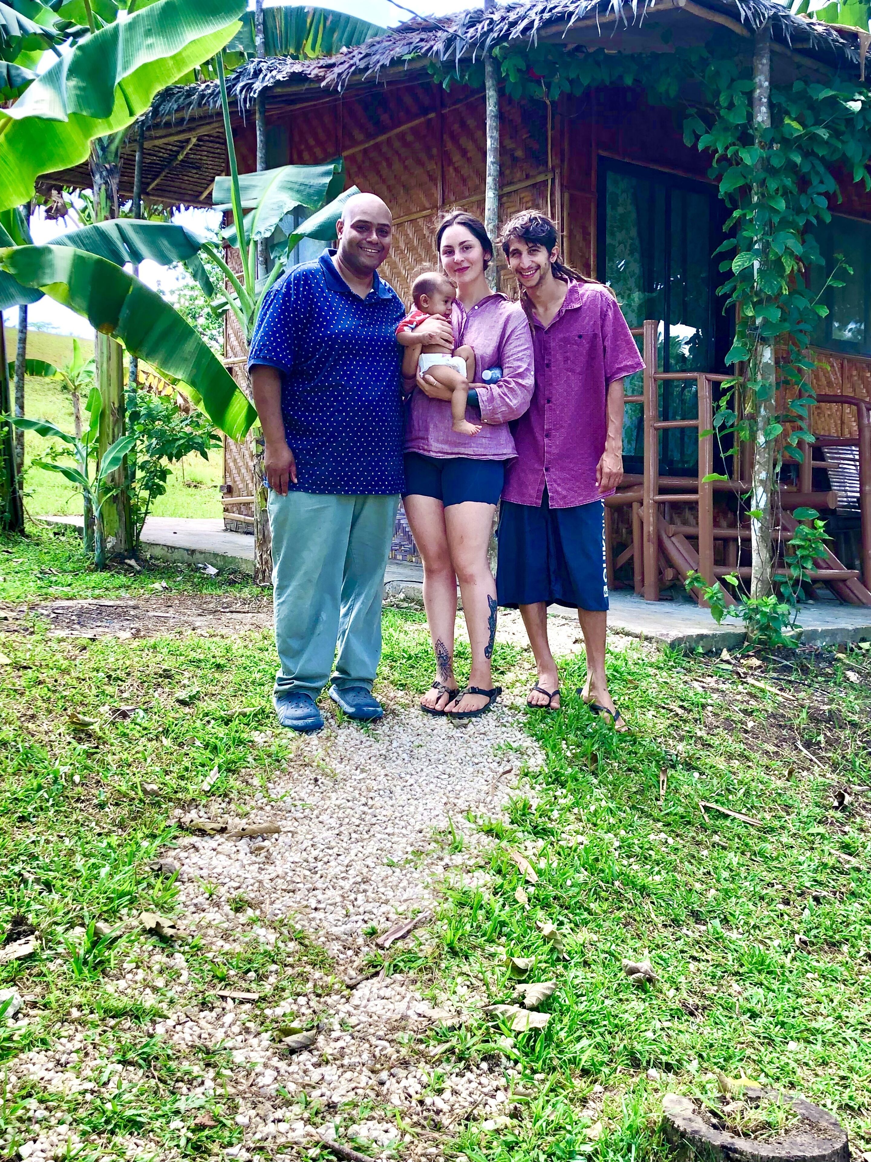 A family of four standing in front of a rustic wooden house with a thatched roof, surrounded by lush greenery.