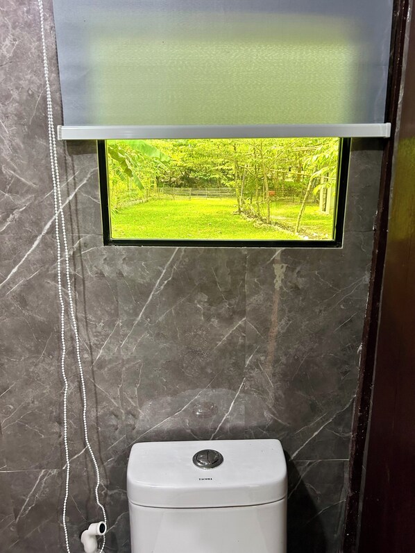 Un inodoro con una pantalla de televisión instalada que muestra una vista de jardín, una pared de mármol y un dispensador de papel higiénico blanco.
