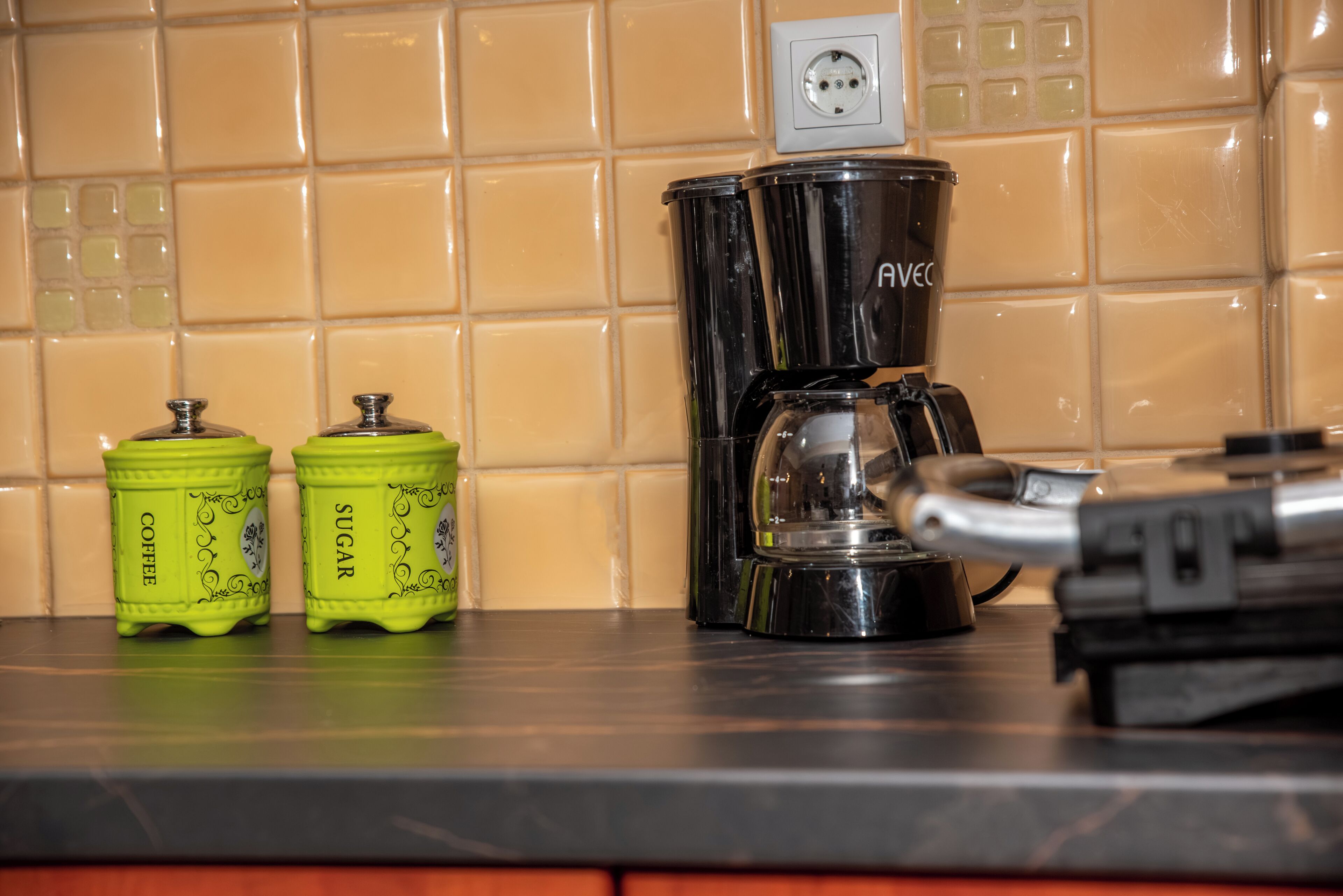 A kitchen counter with a coffee maker, two containers labeled 'Coffee' and 'Sugar,' and a power outlet.