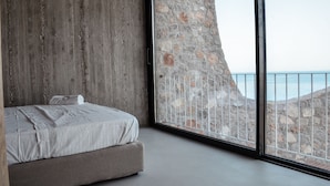 Ein Schlafzimmer mit großem Fenster, das auf eine Steinmauer und den Blick auf das Meer blickt.