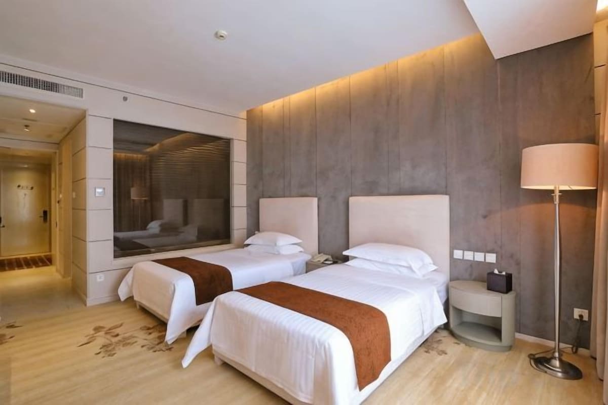A hotel room with two beds, a bedside table, a lamp, and a large window.