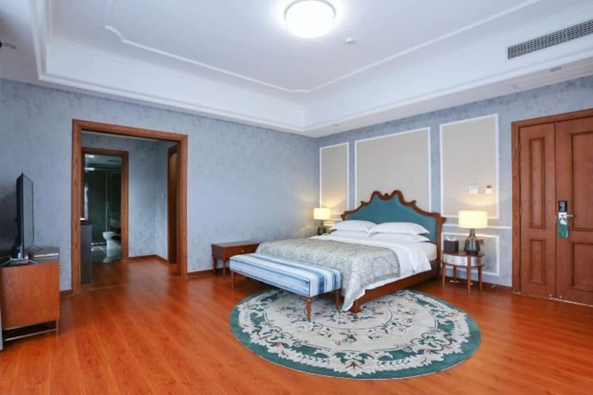A hotel room with a wooden floor, a bed with a blue headboard, two bedside tables with lamps, a large mirror, and a wooden wardrobe.