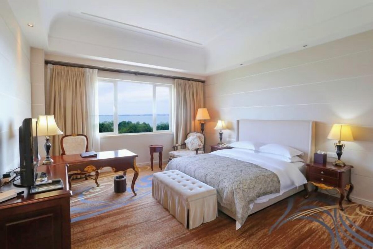 Habitación de hotel con una cama grande, un escritorio, una silla, una mesita, una lámpara y vistas al exterior.