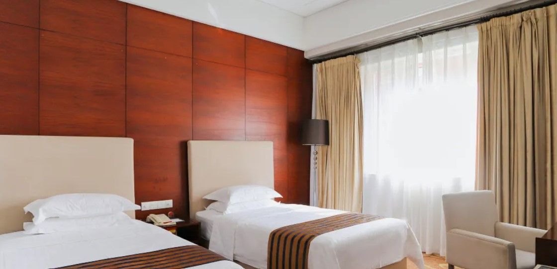 Ein Hotelzimmer mit zwei Betten, einem Schreibtisch, einem Stuhl und einem großen Fenster mit Vorhängen.