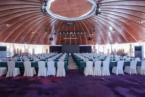 Un salón de conferencias con filas de mesas y sillas, un escenario con iluminación y un techo circular grande.