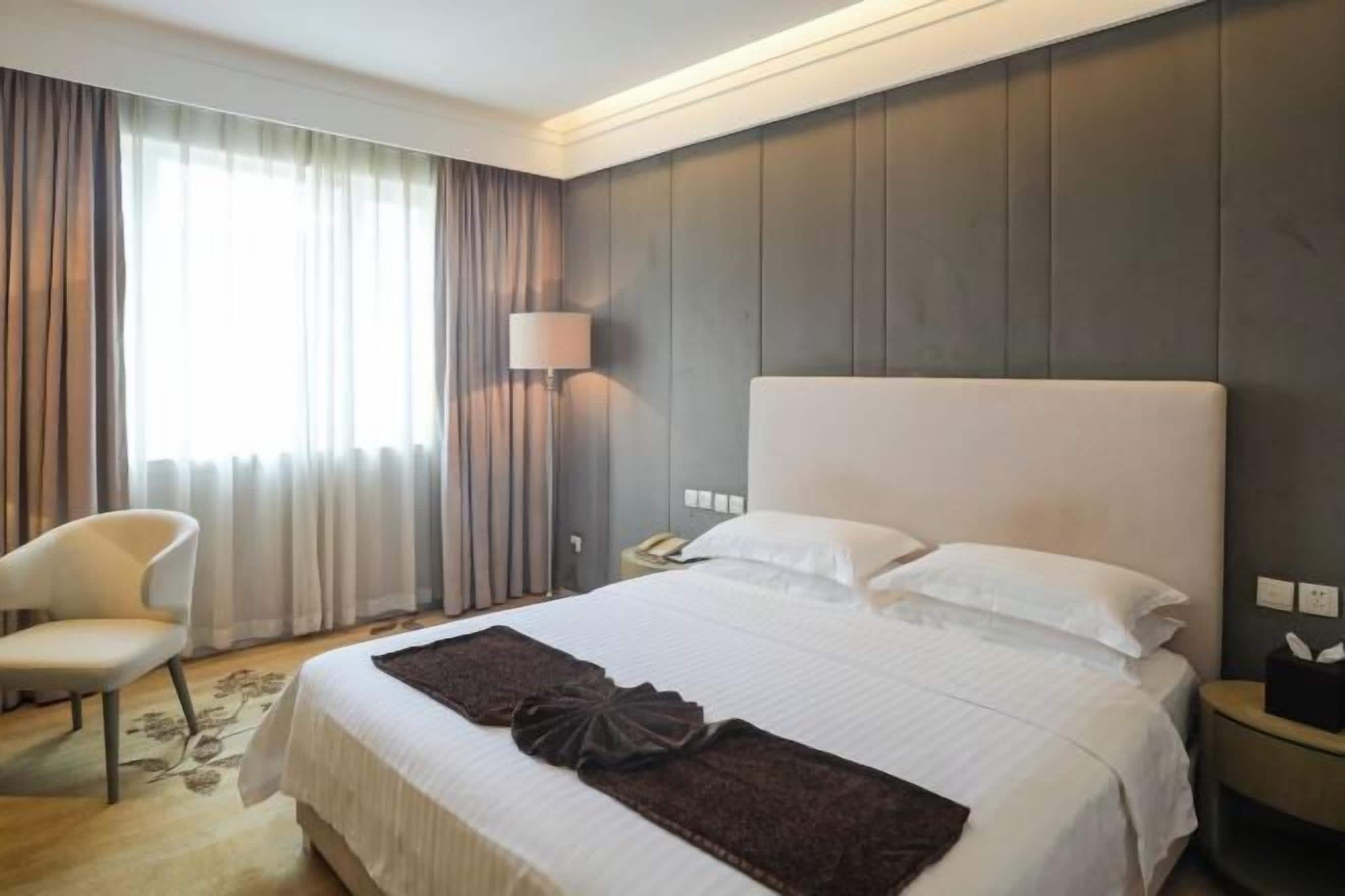 A hotel room with a large bed, a chair, a bedside table with a lamp, and a window with curtains.