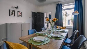 A dining area with a wooden table set for four, chairs, a vase with sunflowers, and a view of the street through a window. - Gateway to the Suffolk Coast (Kessingland)