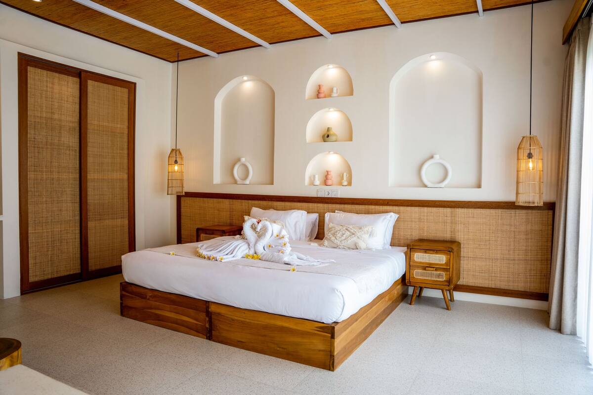 A neatly arranged hotel room with a large bed, wooden headboard, and a wall-mounted shelf with decorative items.