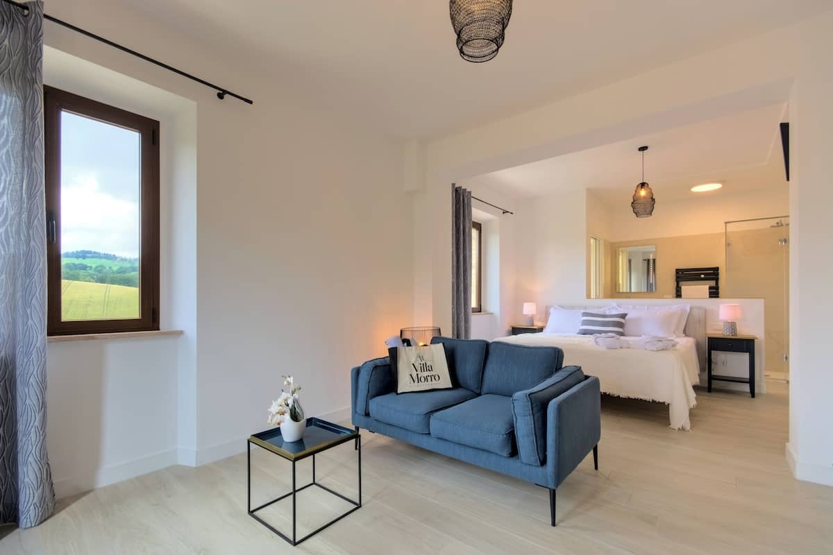 Habitación de hotel moderna con una cama grande, un sofá, una mesita con un florero y vistas al campo.