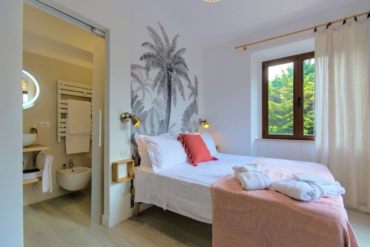 Un dormitorio con cama, mesita de noche, ventana con cortinas, luz en la pared y baño con lavamanos y espejo.