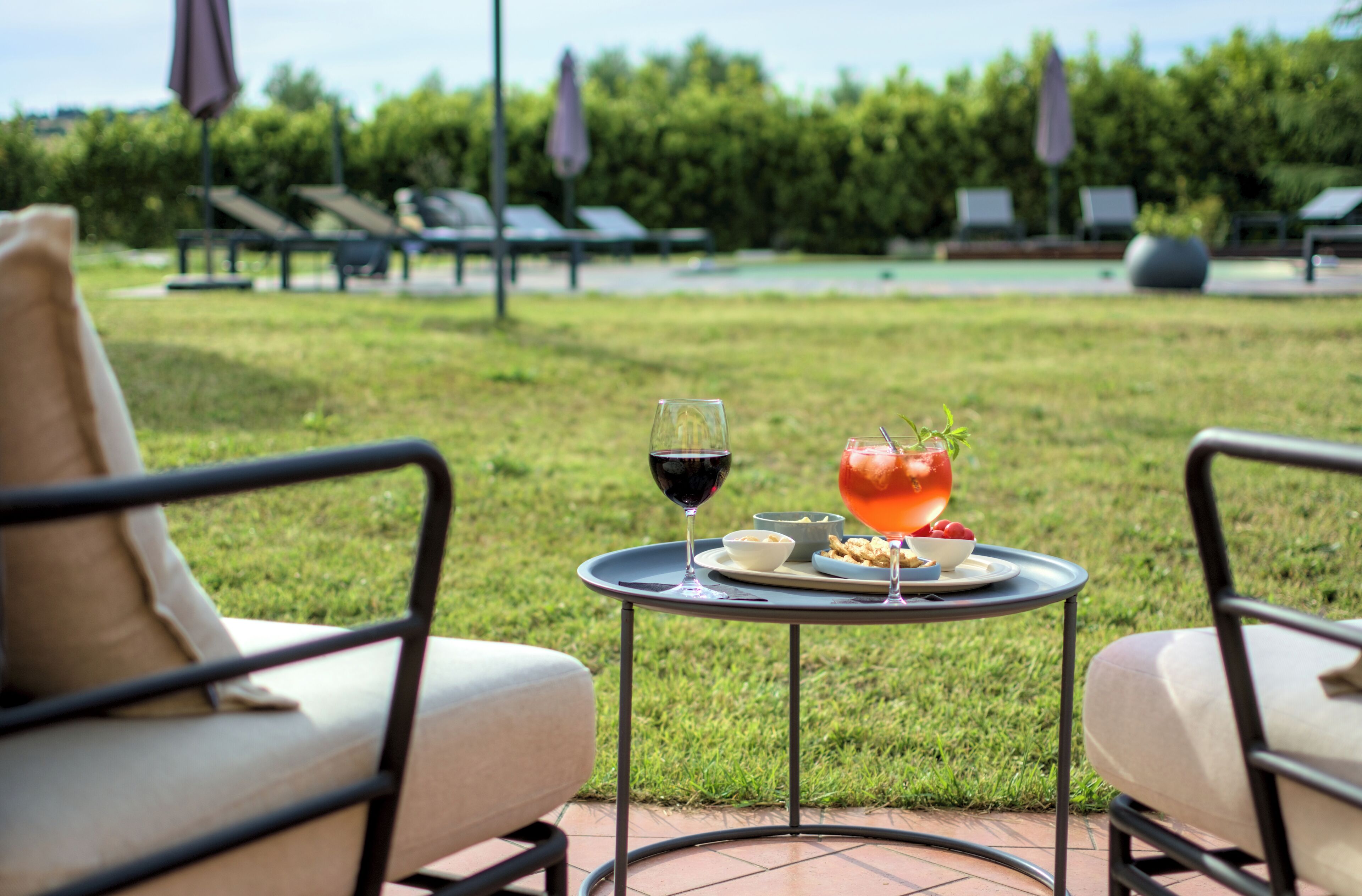 A patio with two chairs, a table with a glass of red wine and a fruit drink, and a pool in the background.
