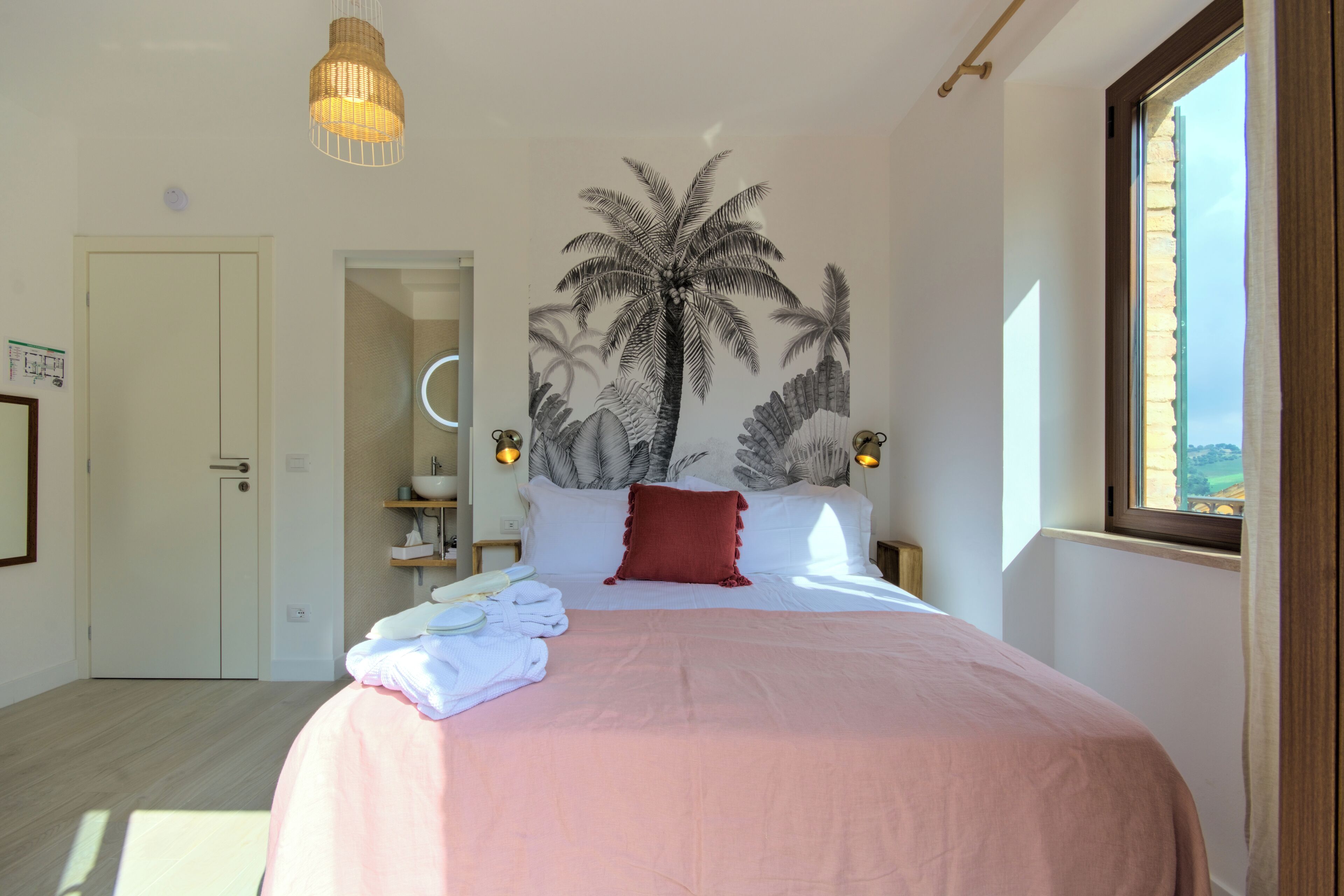A bedroom with a large bed, a wall mural of palm trees, a window with a view, and a door leading to another room.