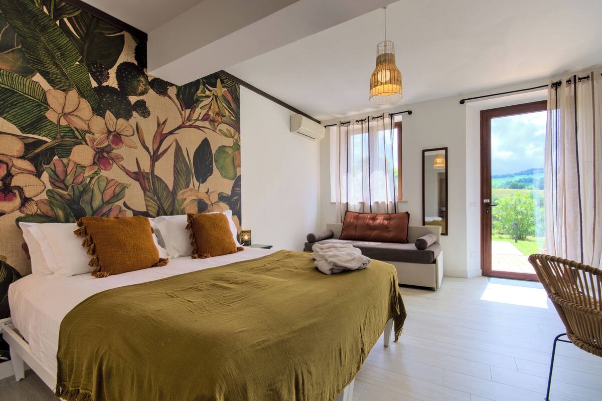 Un dormitorio con una cama grande, un sofá, un sillón de mimbre y vistas a la vegetación a través de la ventana.