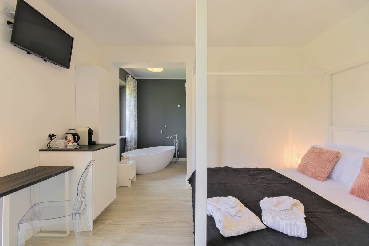 Una habitación de hotel moderna con una televisión de pantalla plana, una zona de cocina con cafetera, una bañera y una cama con toallas.