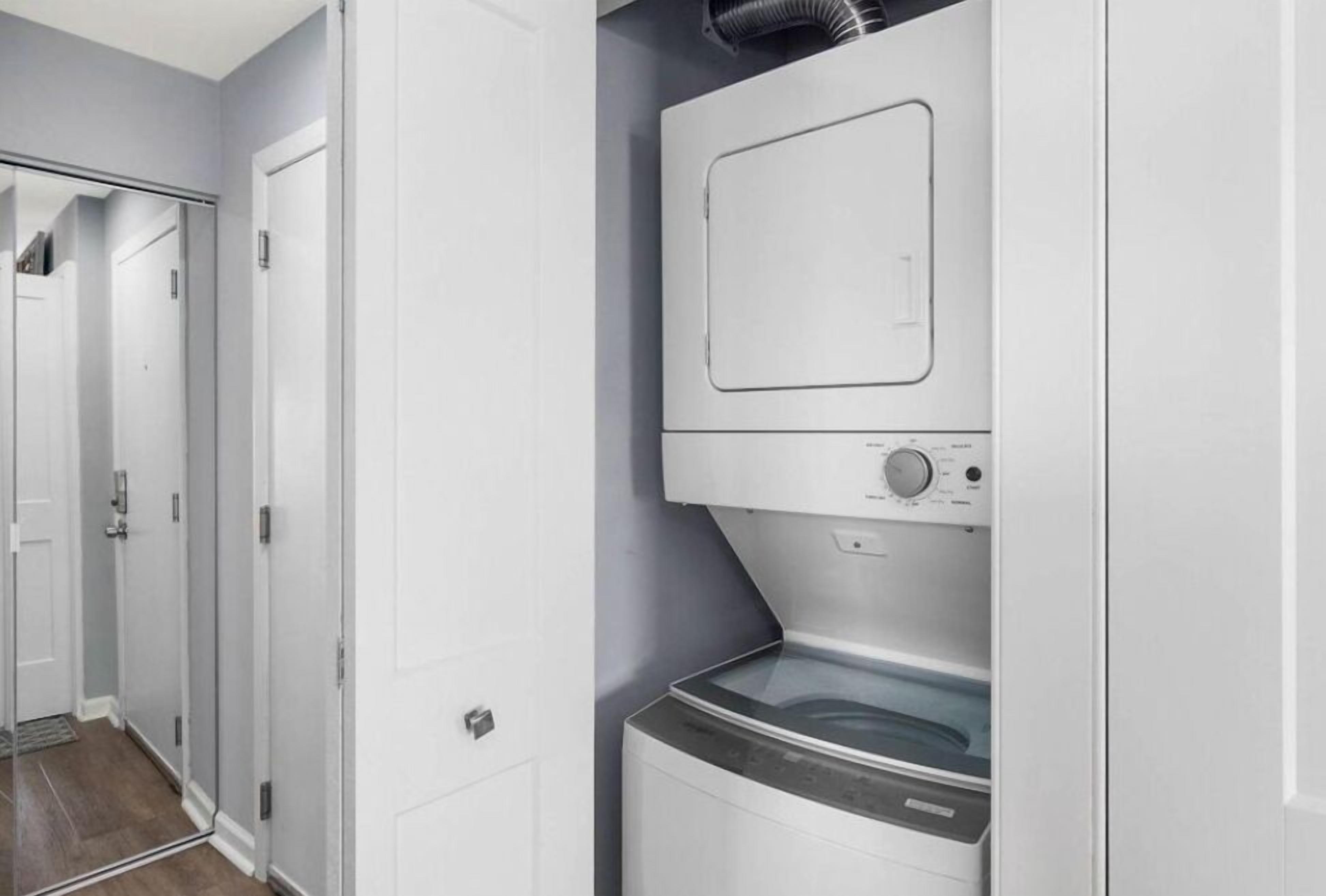 A laundry room with a front-loading washer and dryer, a ventilation system above the dryer, and a door leading to another room.