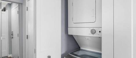 A laundry room with a front-loading washer and dryer, a ventilation system above the dryer, and a door leading to another room.