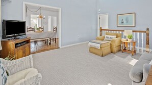 Un salon spacieux avec un téléviseur, un coin repas avec une table à rayures et un coin salon confortable agrémenté d’un coussin orné du mot « RELAX ».