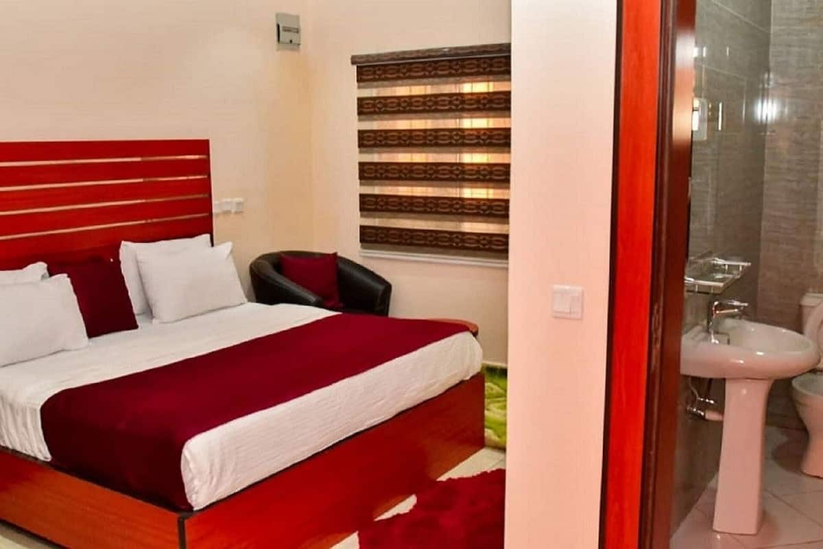 A hotel room with a red bed, white pillows, a black chair, a window with blinds, and a bathroom with a sink and toilet.