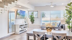A modern living room with a dining area, large windows with a view of the beach, a staircase, and a ceiling fan. - Modern Beachfront Retreat 2 King Suites + (Puerto Peñasco)