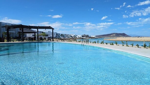 A clear blue pool with lounge chairs and a pergola, surrounded by buildings and a beach. - Modern Beachfront Retreat 2 King Suites + (Puerto Peñasco)