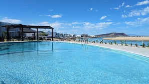 A clear blue pool with lounge chairs and a pergola, surrounded by buildings and a beach.