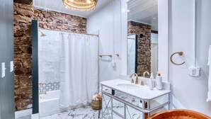 A bathroom with a stone wall, a bathtub, a white sink with gold fixtures, and a mirror. - Robin Ave. #9 (Mason)
