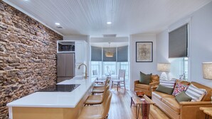 A modern kitchen with a stone wall, a central island with bar stools, a dining area with a table and chairs, and a living area with a sofa and cushions. - Robin Ave. #9 (Mason)