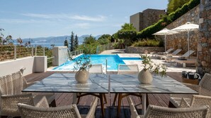 Zona junto a la piscina con mesa y sillas, pared de piedra y vista al mar.