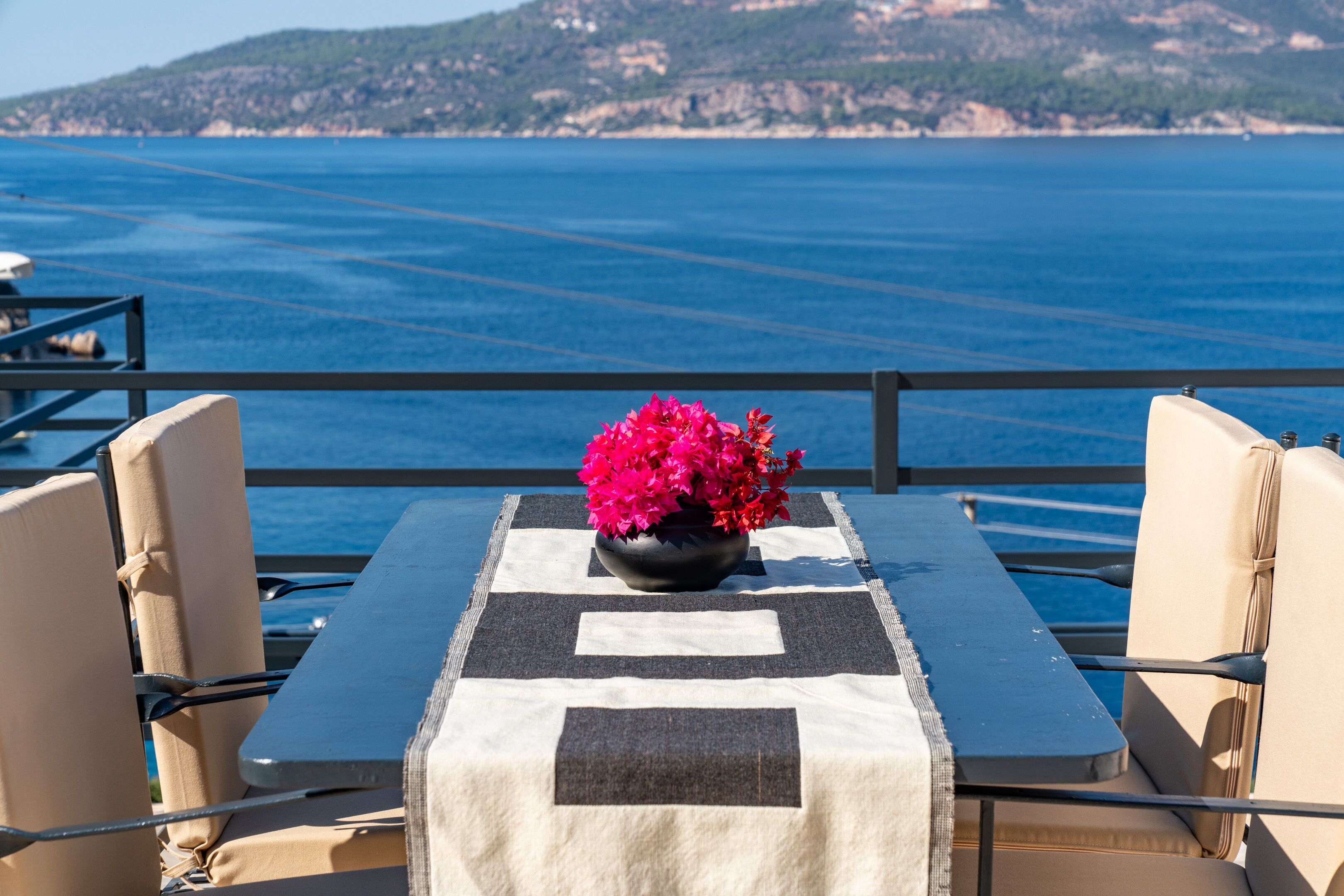Una mesa con mantel blanco y negro, dos sillas color beige y un florero negro con flores rosadas.