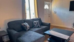 Ein Wohnzimmer mit grauem Sofa, zwei gemusterten Kissen, einem Couchtisch und einer Tür im Hintergrund.