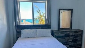 Ein Schlafzimmer mit einem Bett, einem Fenster mit Blick auf den Strand und einer Holzkommode.