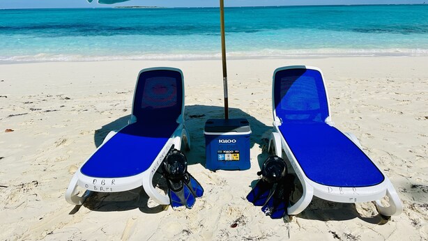 Zwei blaue Liegest ühle mit einem blauen Sonnenschirm und einer Kühlbox mit dem Aufdruck "IGLOO" an einem Sandstrand.
