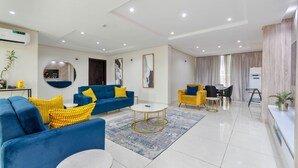 Uma sala de estar moderna com um sofá azul, almofadas amarelas, uma mesa de centro redonda e uma área de jantar com cadeiras amarelas.