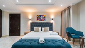 Quarto de hotel moderno com uma cama grande, cabeceira estofada, mesa de cabeceira, uma cadeira e um quadro na parede.