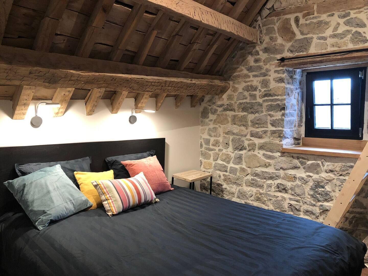 Een slaapkamer met een stenen muur, een houten plafond, een bed met veel kussens en een kleine tafel.