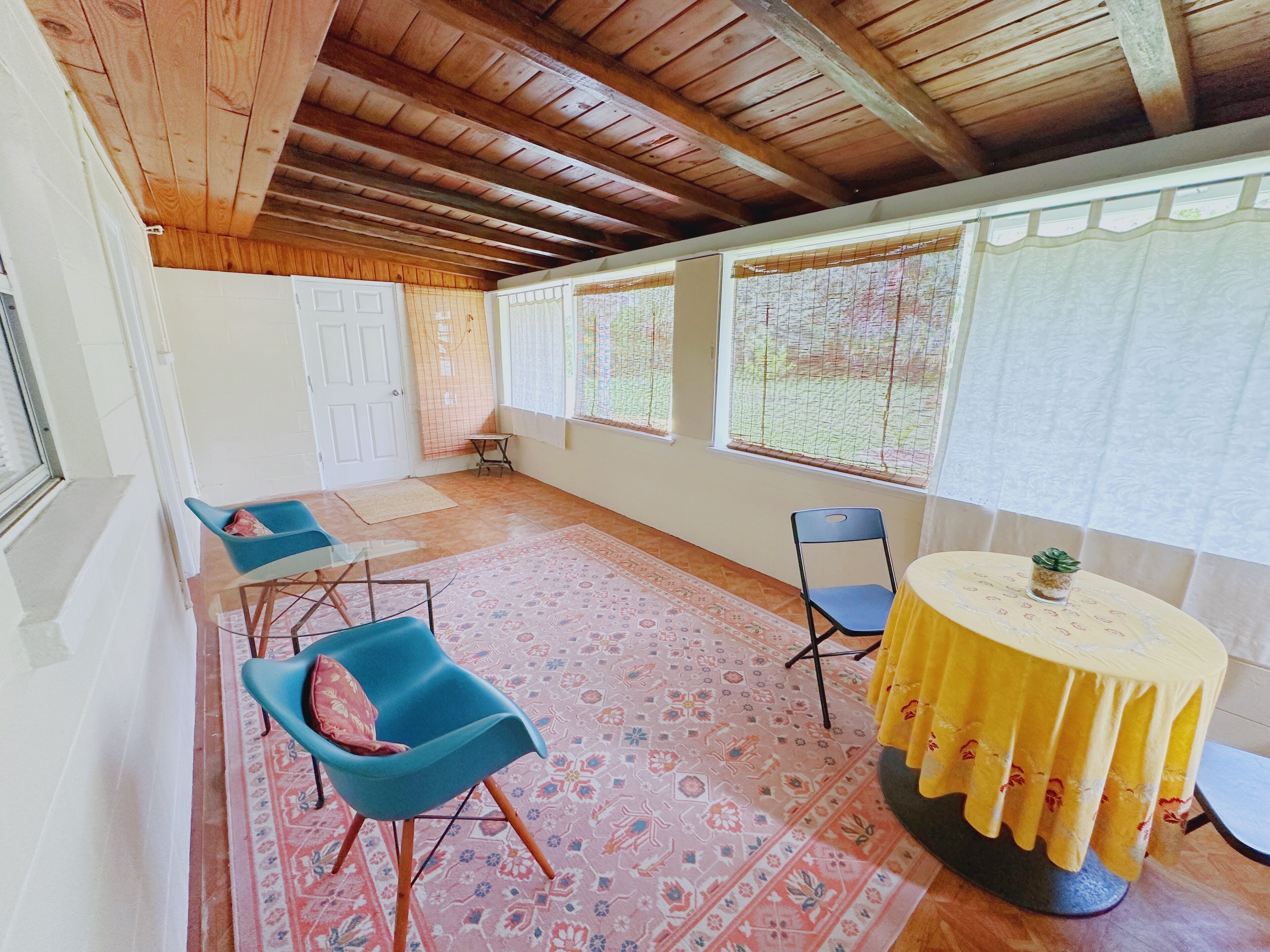 A room with a wooden ceiling, a patterned rug, a blue chair, a yellow table, and a window with a curtain.