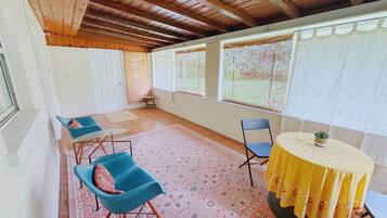 A room with a wooden ceiling, a patterned rug, a blue chair, a yellow table, and a window with a curtain.