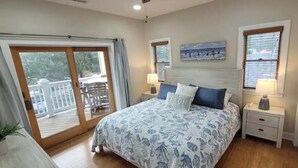 A bedroom with a large bed, a nightstand, a window with blinds, and a view of the outdoors.