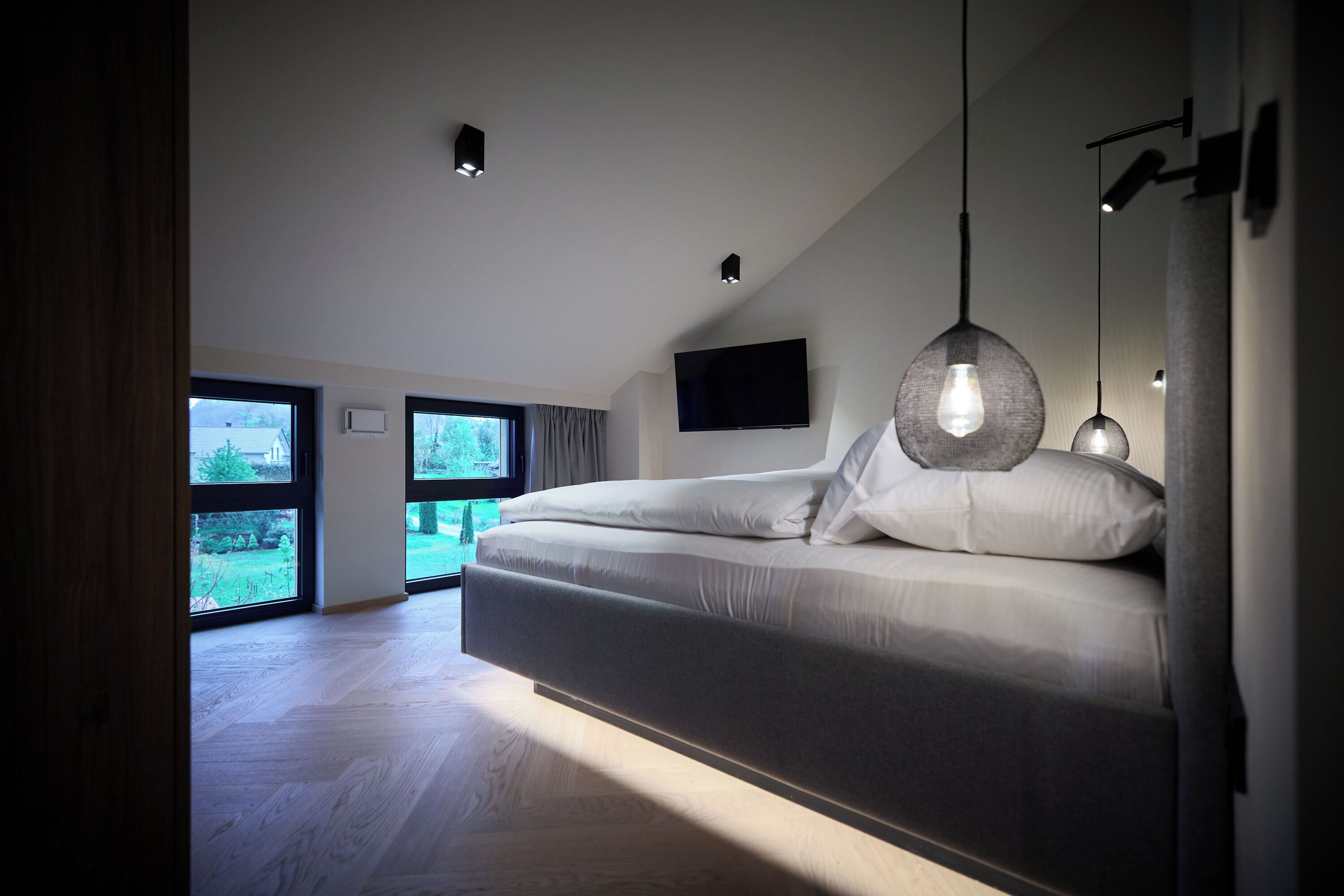 A modern bedroom with a large bed, a flat-screen TV mounted on the wall, and a view of the outdoors through large windows.