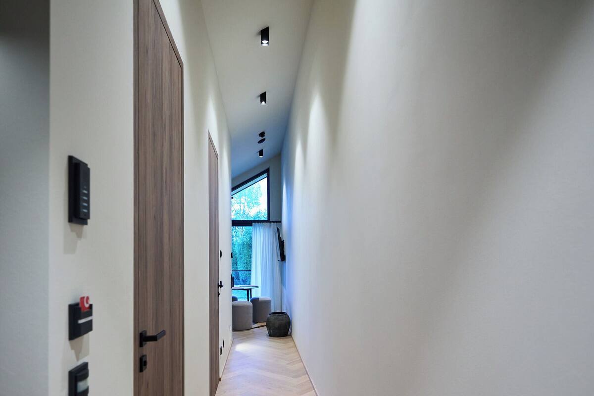 A modern hallway with wooden flooring and white walls, leading to a room with a large window and a view of greenery.