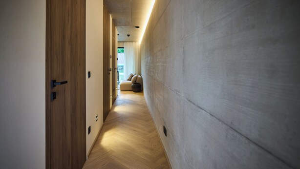 A hallway with wooden flooring and a concrete wall, leading to a seating area with cushions.