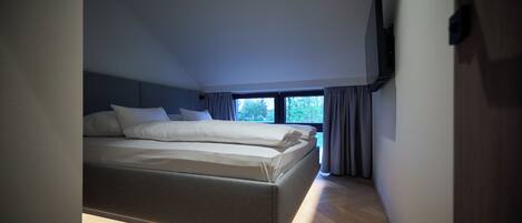 A modern bedroom with a bed, a TV mounted on the wall, and a window with curtains.