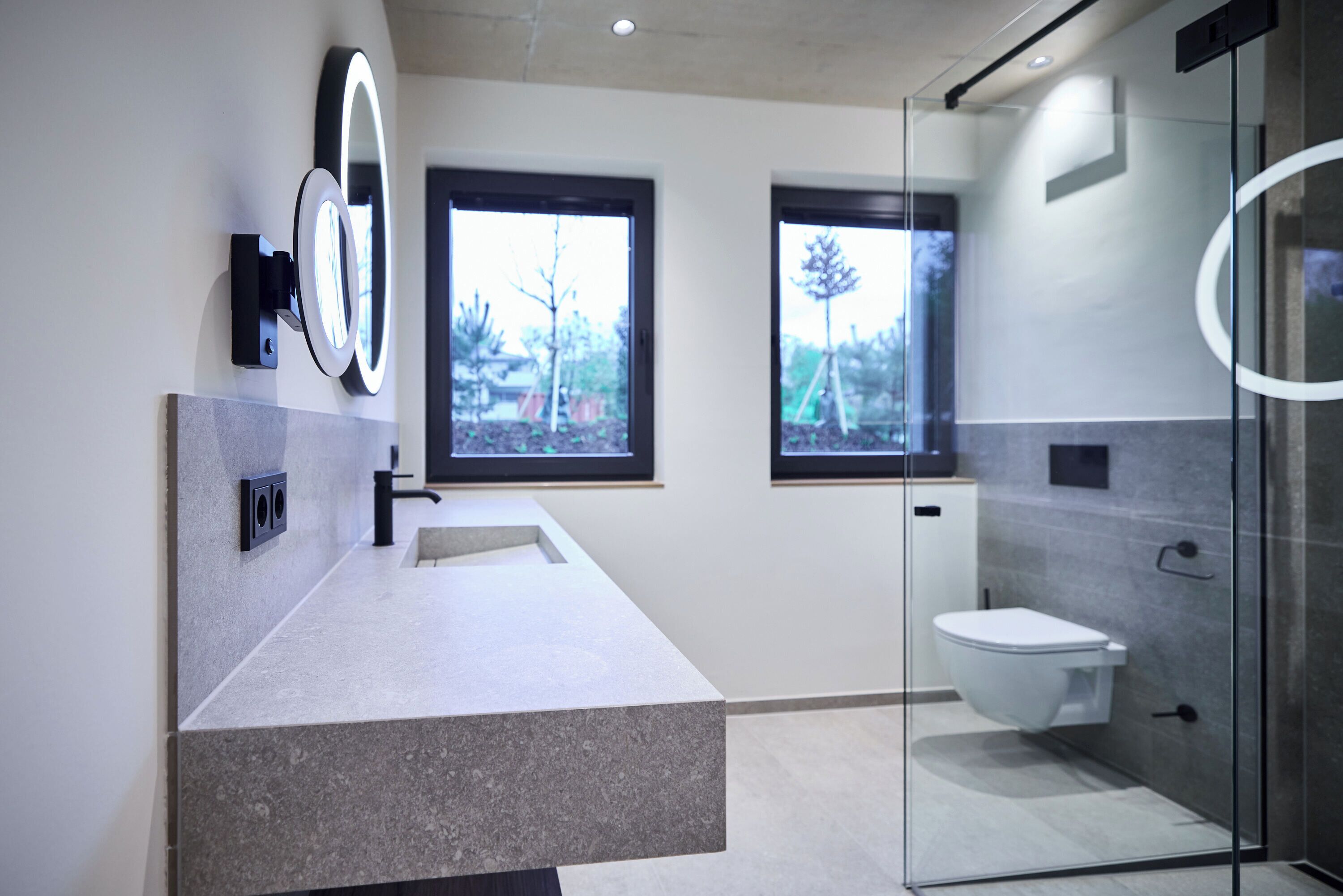 Une salle de bain moderne dotée d’un comptoir en granit, d’une cabine de douche en verre et de deux fenêtres qui laissent entrer la lumière naturelle.
