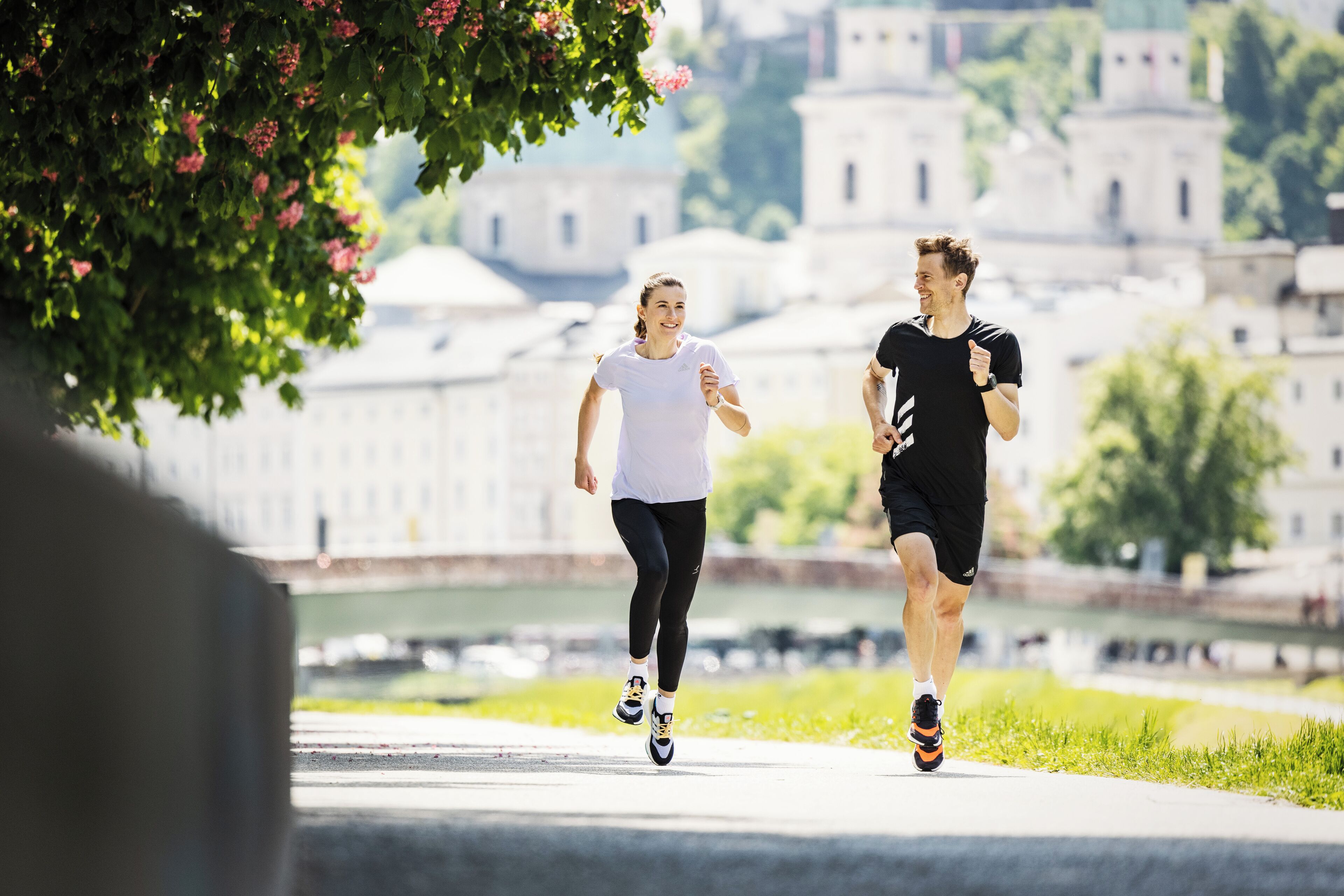 Twee personen joggen op een pad, met een rivier en gebouwen op de achtergrond.