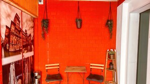 Um quarto com parede de tijolos vermelhos, duas bancos de madeira, duas cadeiras pretas, uma mesinha, uma lixeira e plantas penduradas.