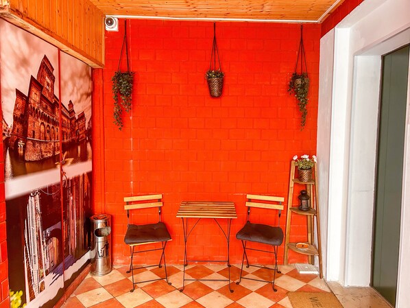 A room with a red brick wall, two wooden benches, two black chairs, a small table, a trash can, and hanging plants.