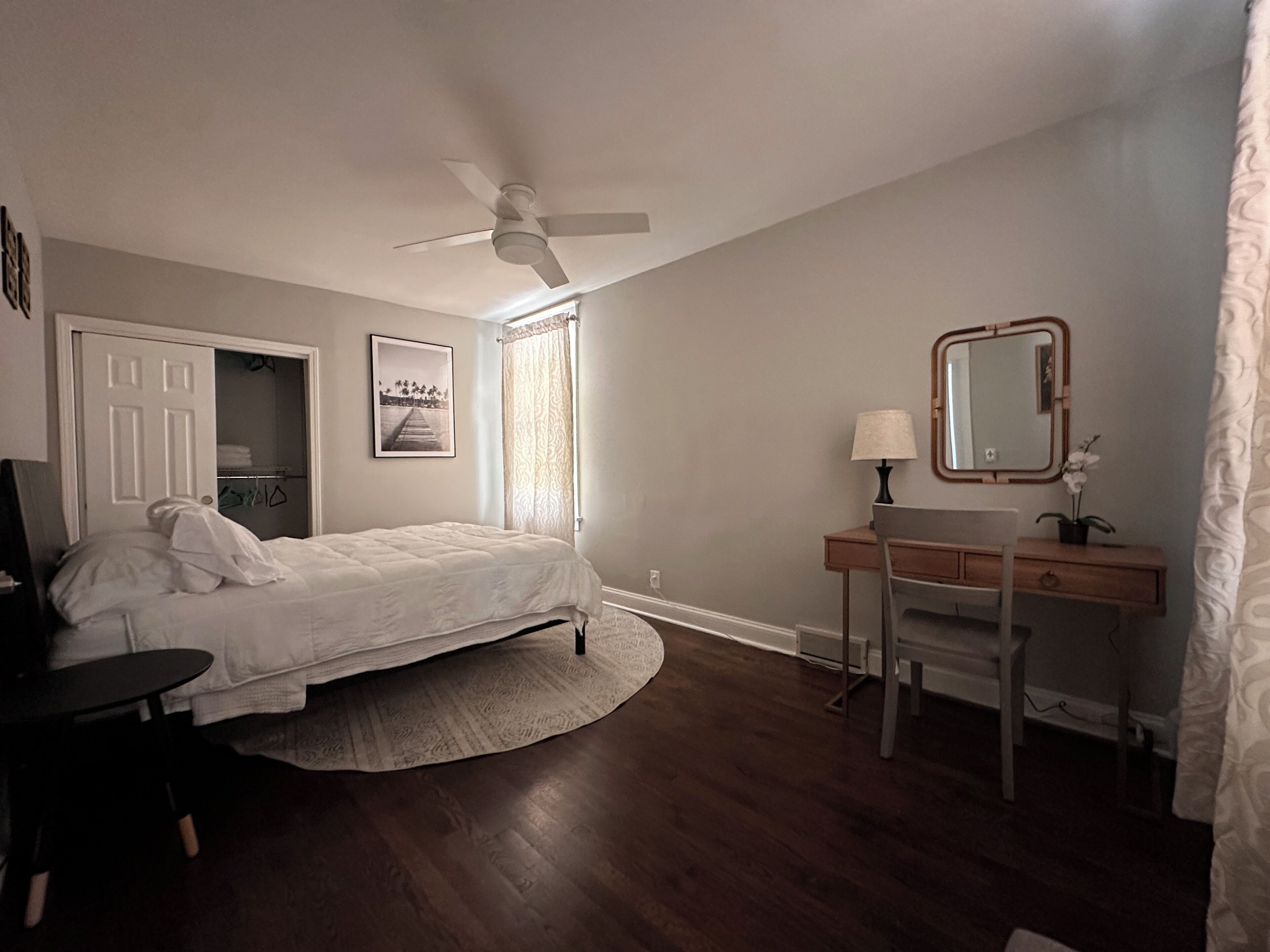 A bedroom with a bed, a desk, a chair, a ceiling fan, a window with curtains, and a framed picture on the wall.