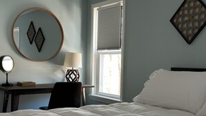 A bedroom with a bed, a desk, a chair, a lamp, a mirror, and a window with blinds. - Cozy apartment right in Downtown Downers Grove (Downers Grove)