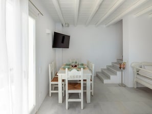 Un área de comedor con una mesa dispuesta para cuatro, sillas blancas, un televisor montado en la pared y una escalera que sube a otro nivel.