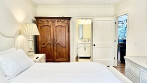 Une chambre à coucher avec un grand lit, une armoire en bois, une table de chevet avec une lampe, une salle de bain avec un miroir et un ventilateur de plafond.